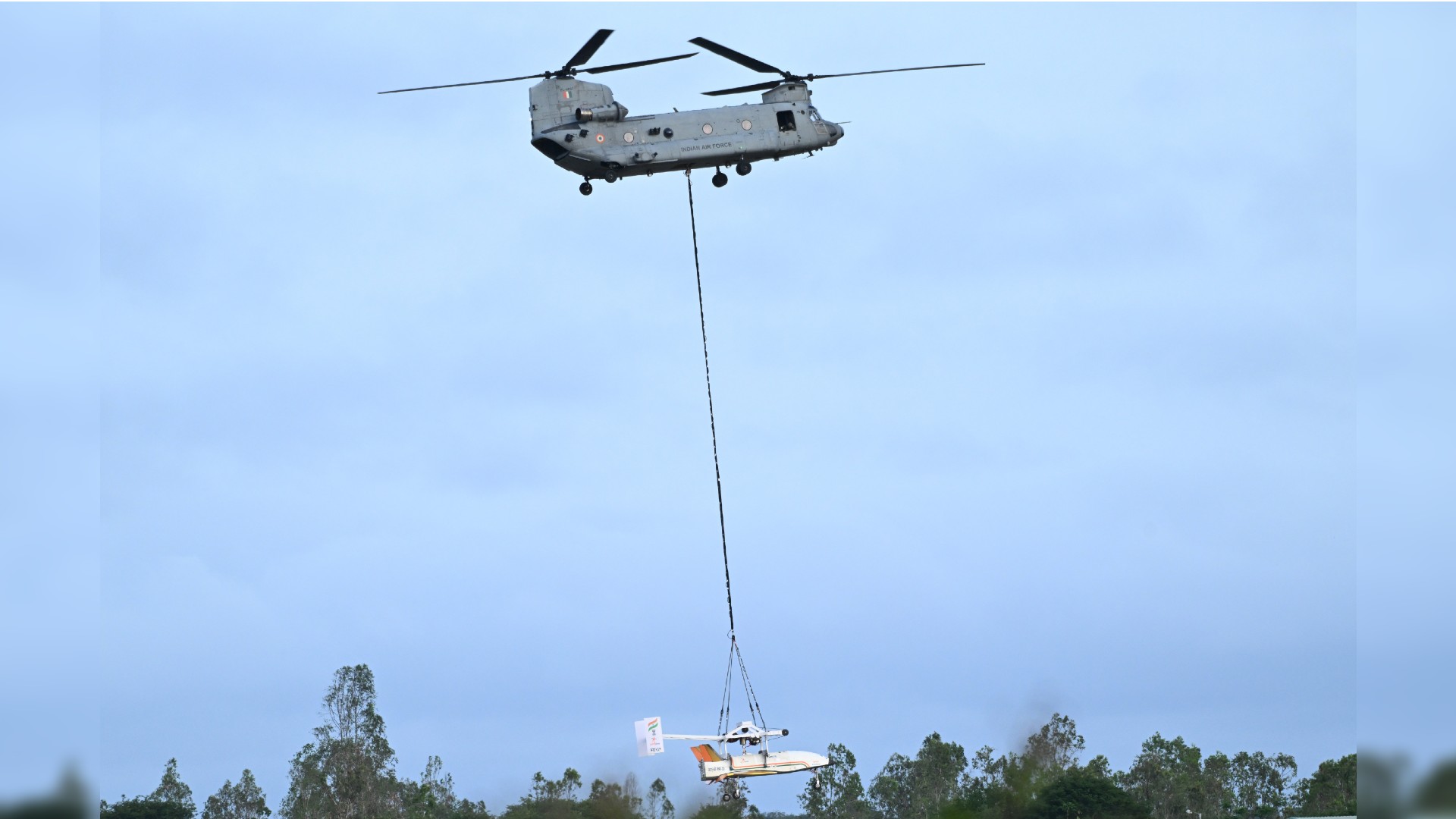 یک هلیکوپتر بزرگ دو روتور هواپیمای سفید و بدون پنجره را در انتهای یک کابل بلند در زیر خود حمل می کند.